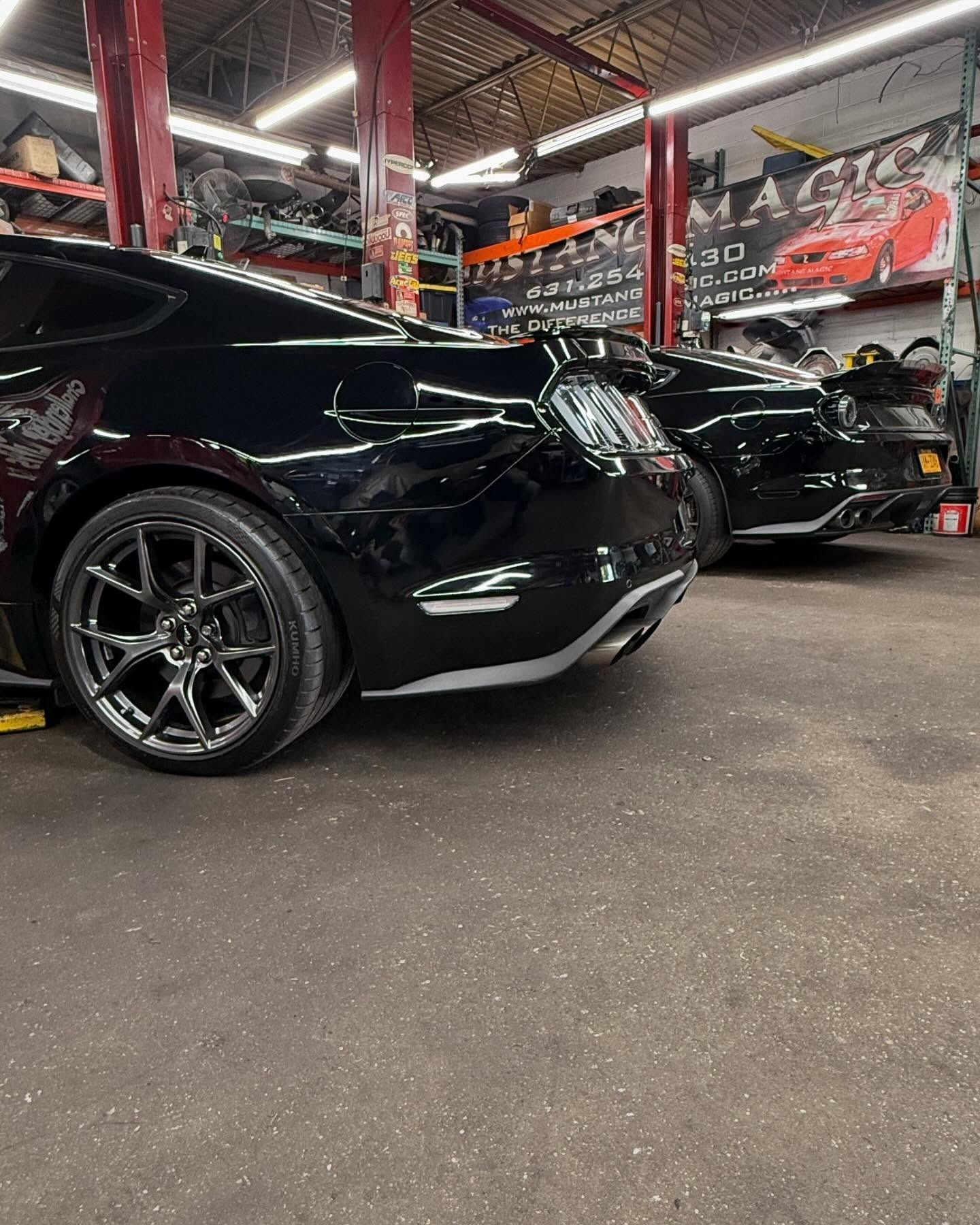 Two black Mustangs parked inside a garage. One has chrome wheels.