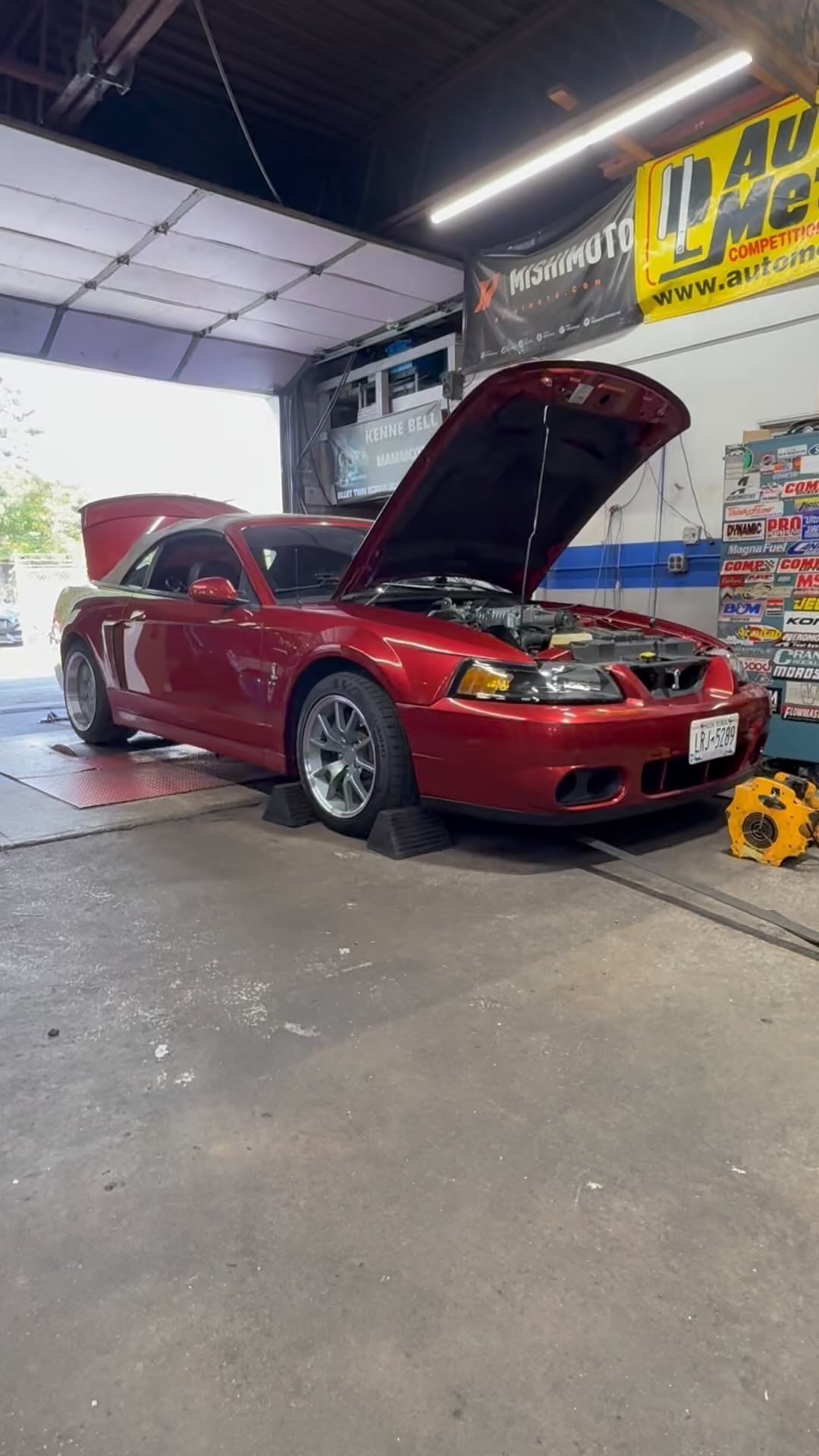 Red Ford Mustang with the hood open in a garage, possibly undergoing repair.