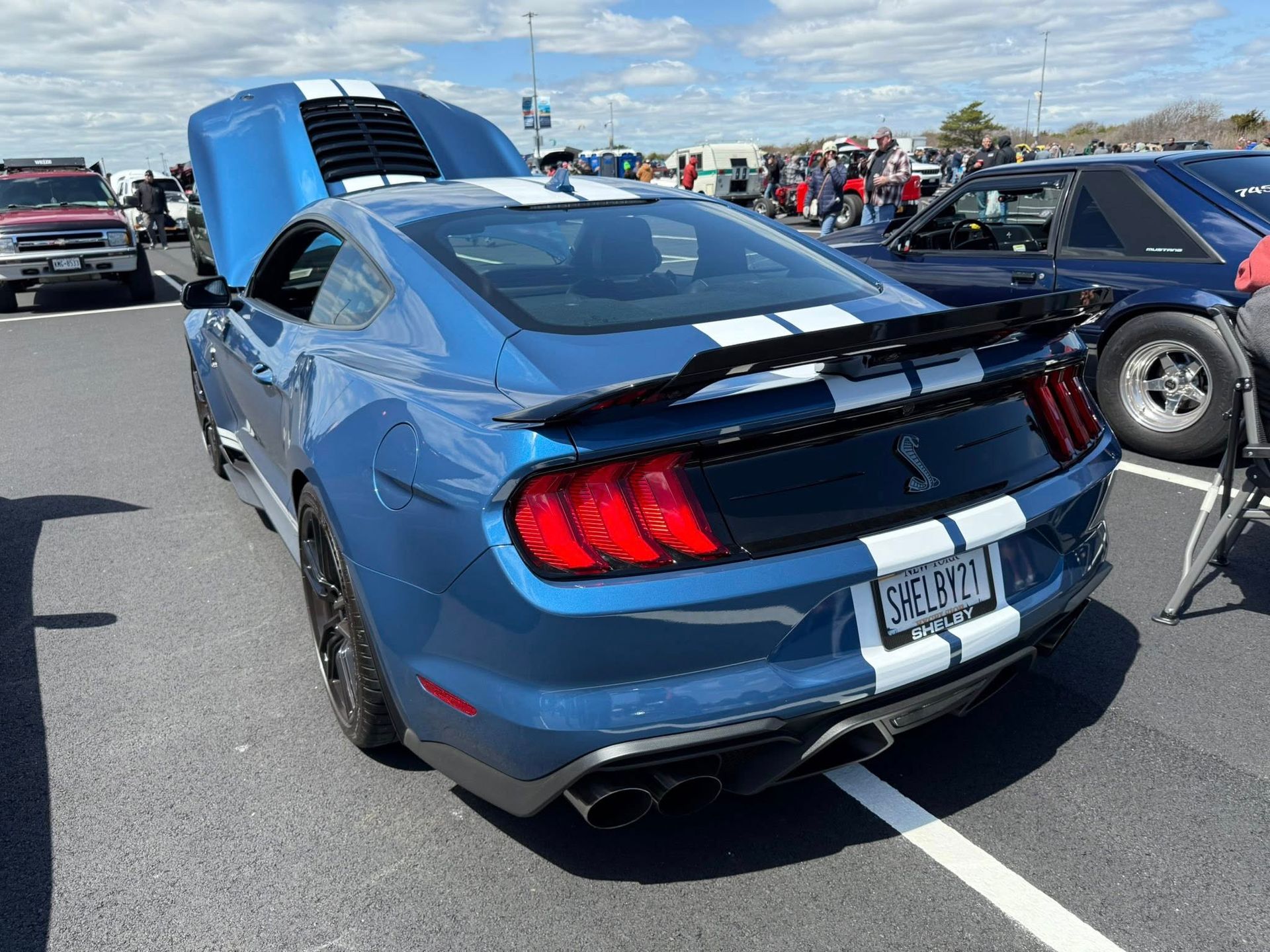 Blue Shelby Mustang with white stripes, hood open, parked outdoors at a car show.