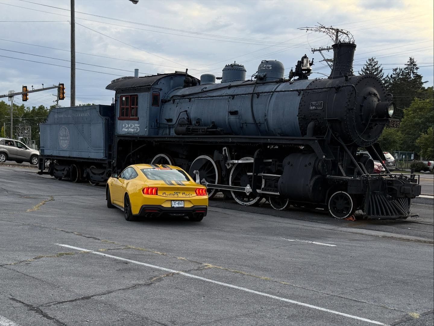 Yellow car on road next to large, dark blue steam engine train.