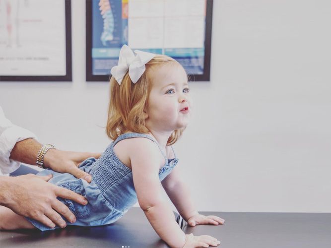 A young girl with a white bow, in a blue dress, is on a table at a doctor's office while being checked by a medical professional.