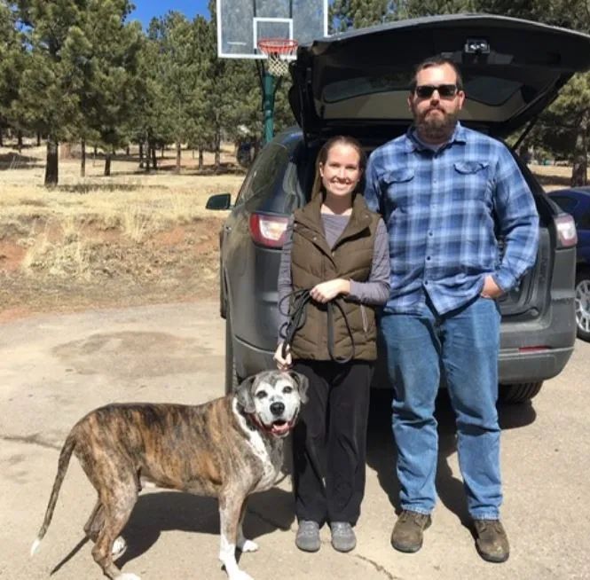 A man and woman standing next to a dog in front of a car