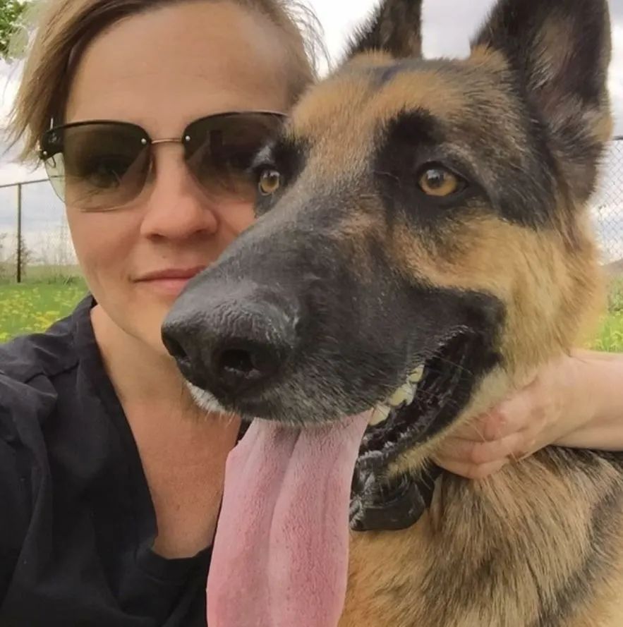 A woman wearing sunglasses holds a german shepherd with its tongue hanging out
