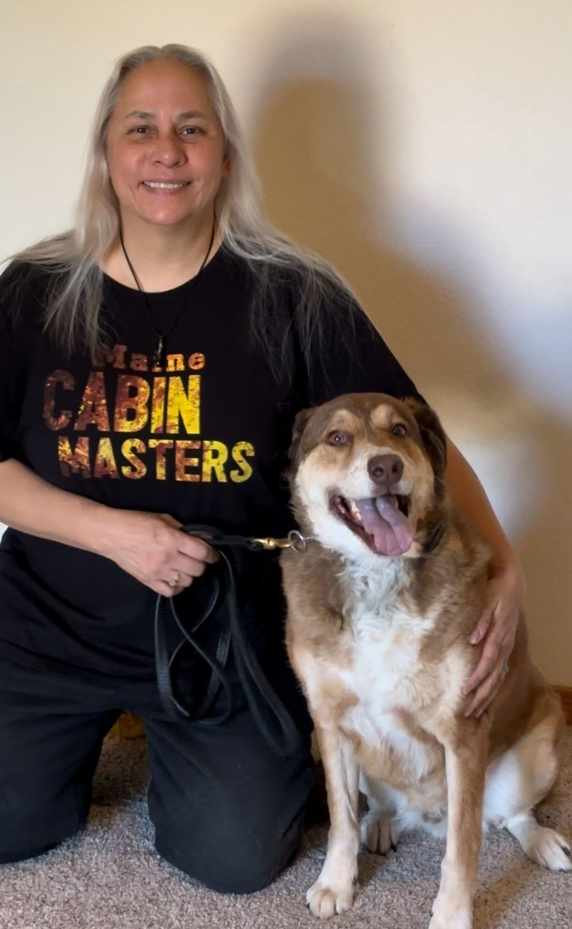 A woman is kneeling down next to a dog wearing a cabin masters shirt.