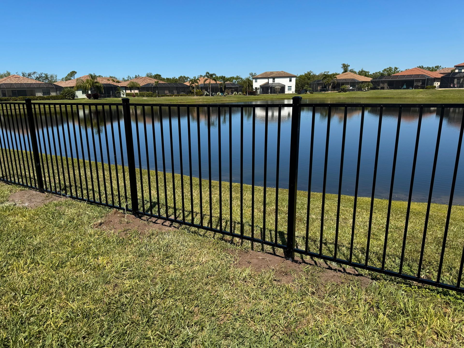 A black fence bordering a lake, green grass in the foreground, and houses in the background under a blue sky.