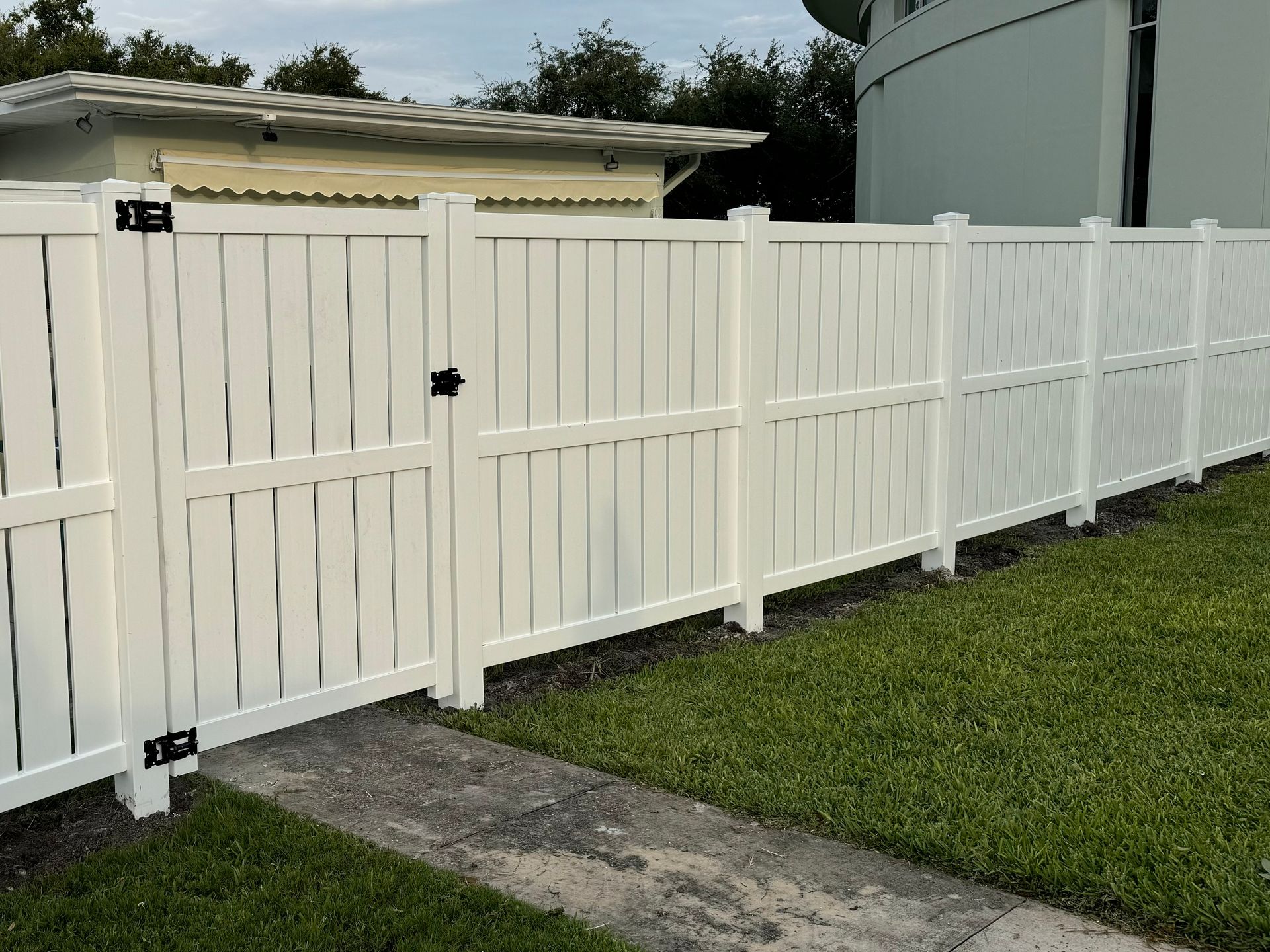 White vinyl fence with gate, on green grass, next to a paved pathway.