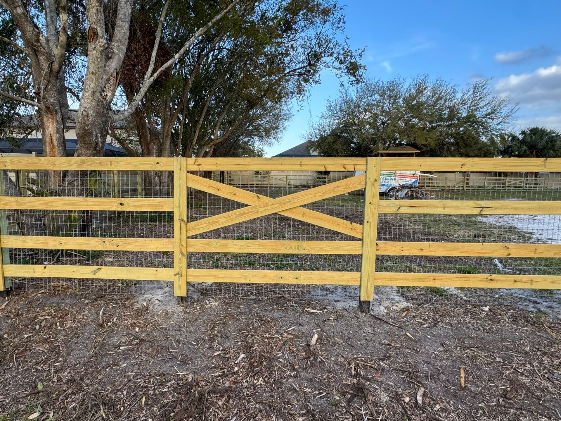 Wooden fence with gate, set against a tree and a partly cloudy sky. The gate features an 