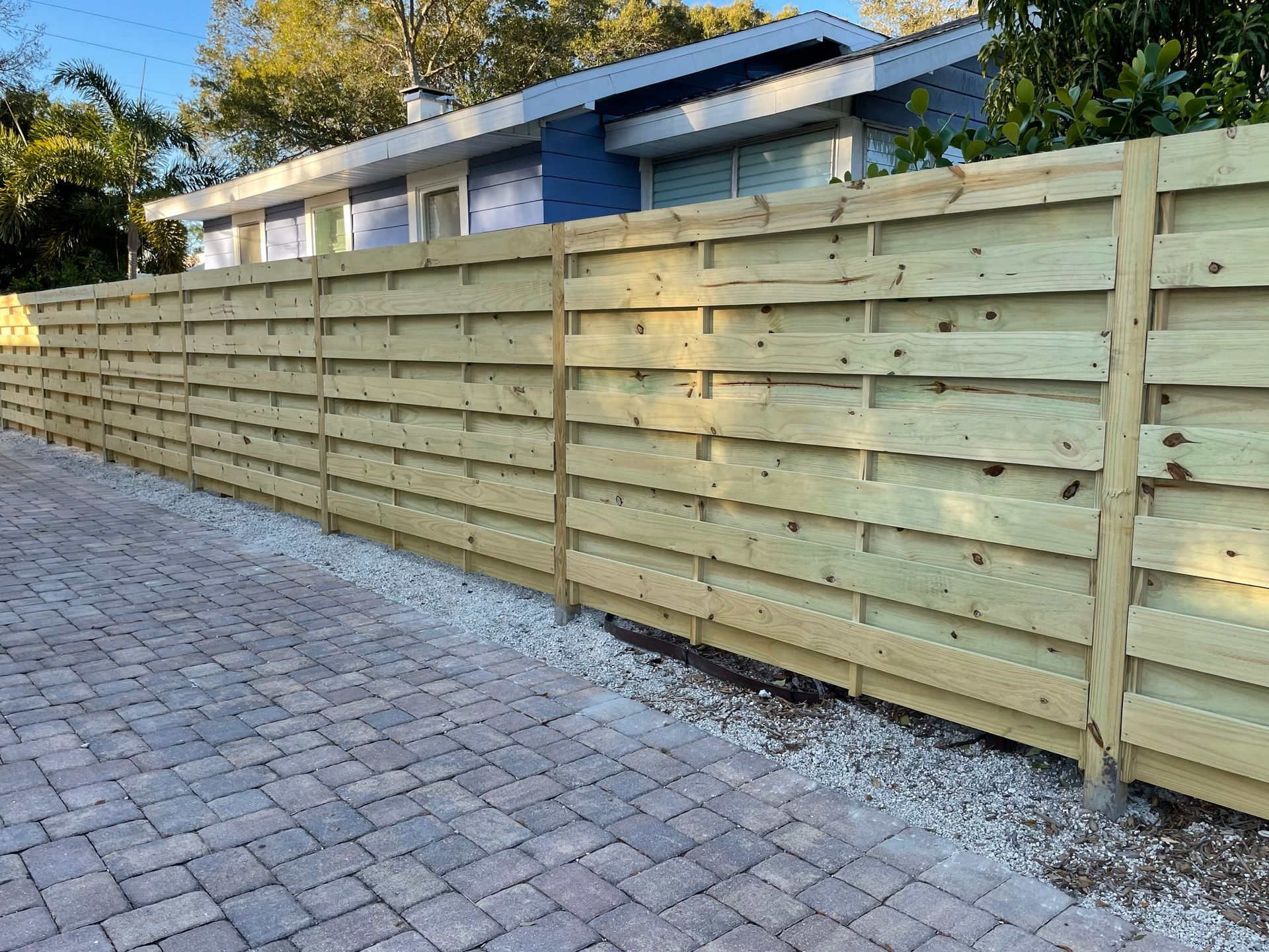 A wooden horizontal slat fence in front of a house, set on gravel, with a brick paver driveway.