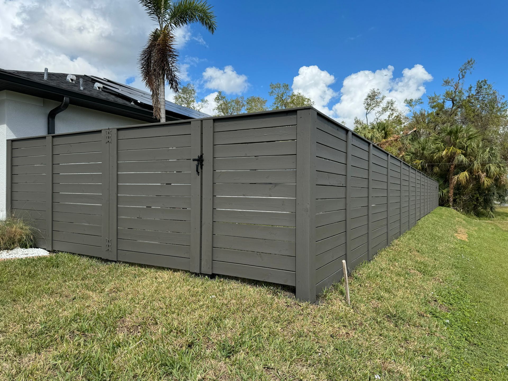 Gray horizontal slat fence with gate, set against a lawn and blue sky with clouds.