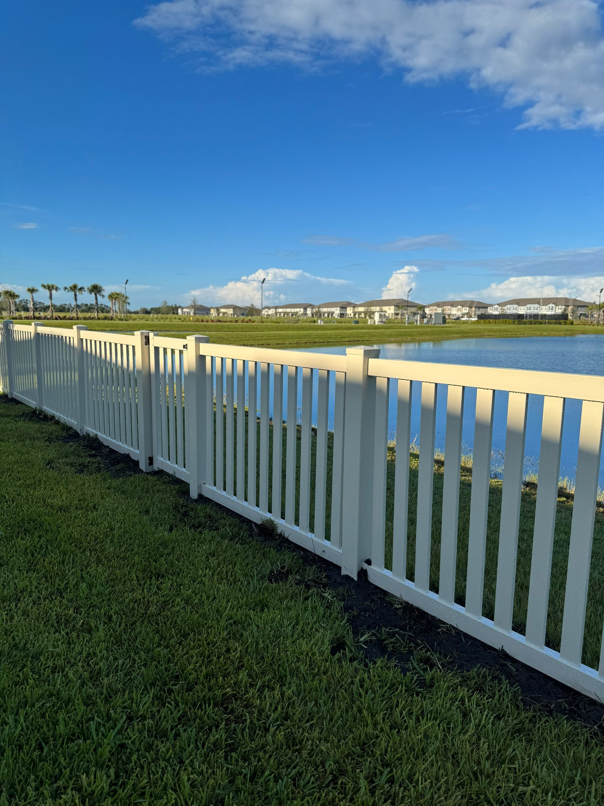 A white picket fence borders a grassy area and a lake under a blue sky with clouds.