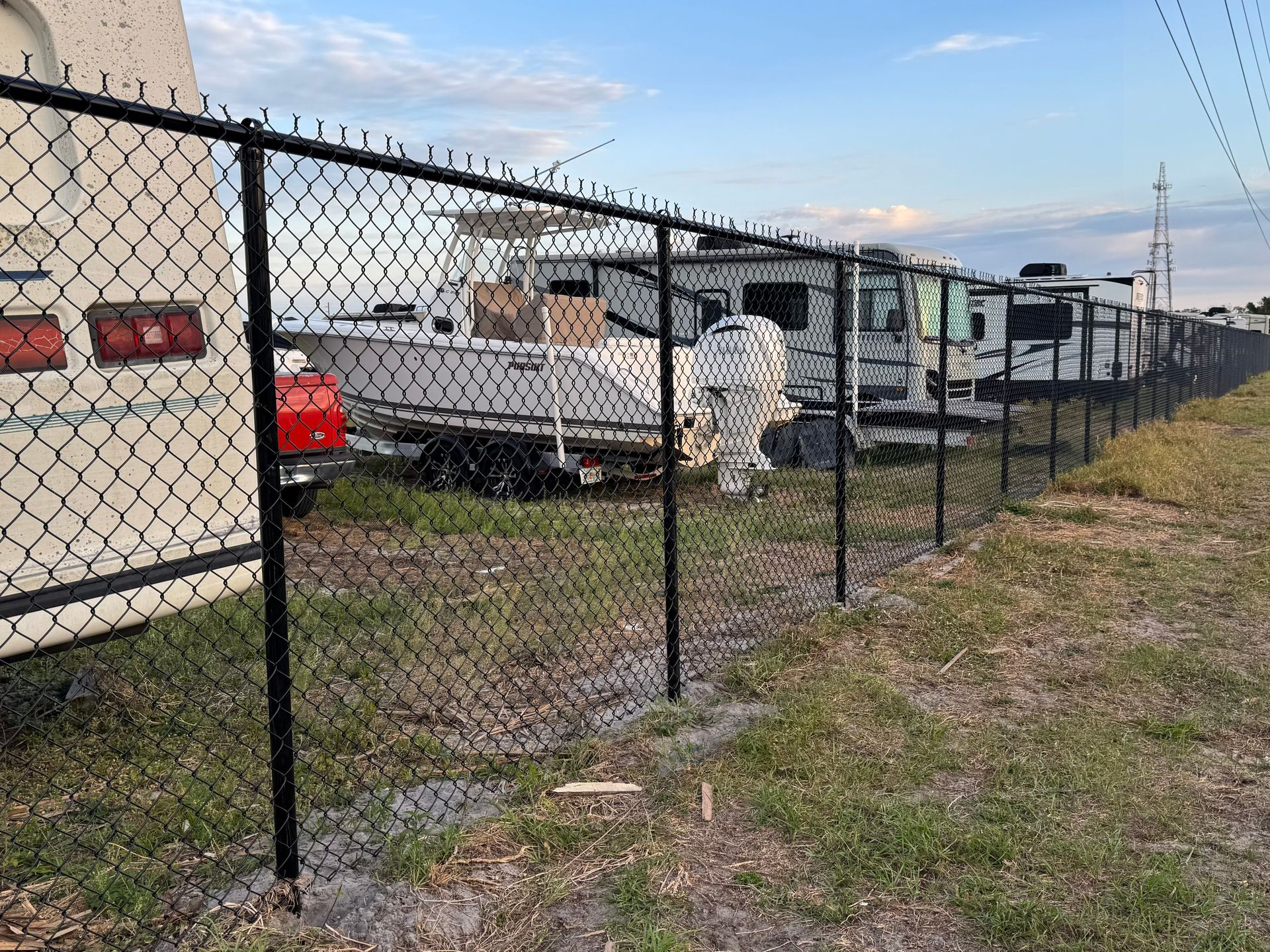 Black chain-link fence bordering a grassy area with RVs and a boat parked behind it, under a cloudy sky.