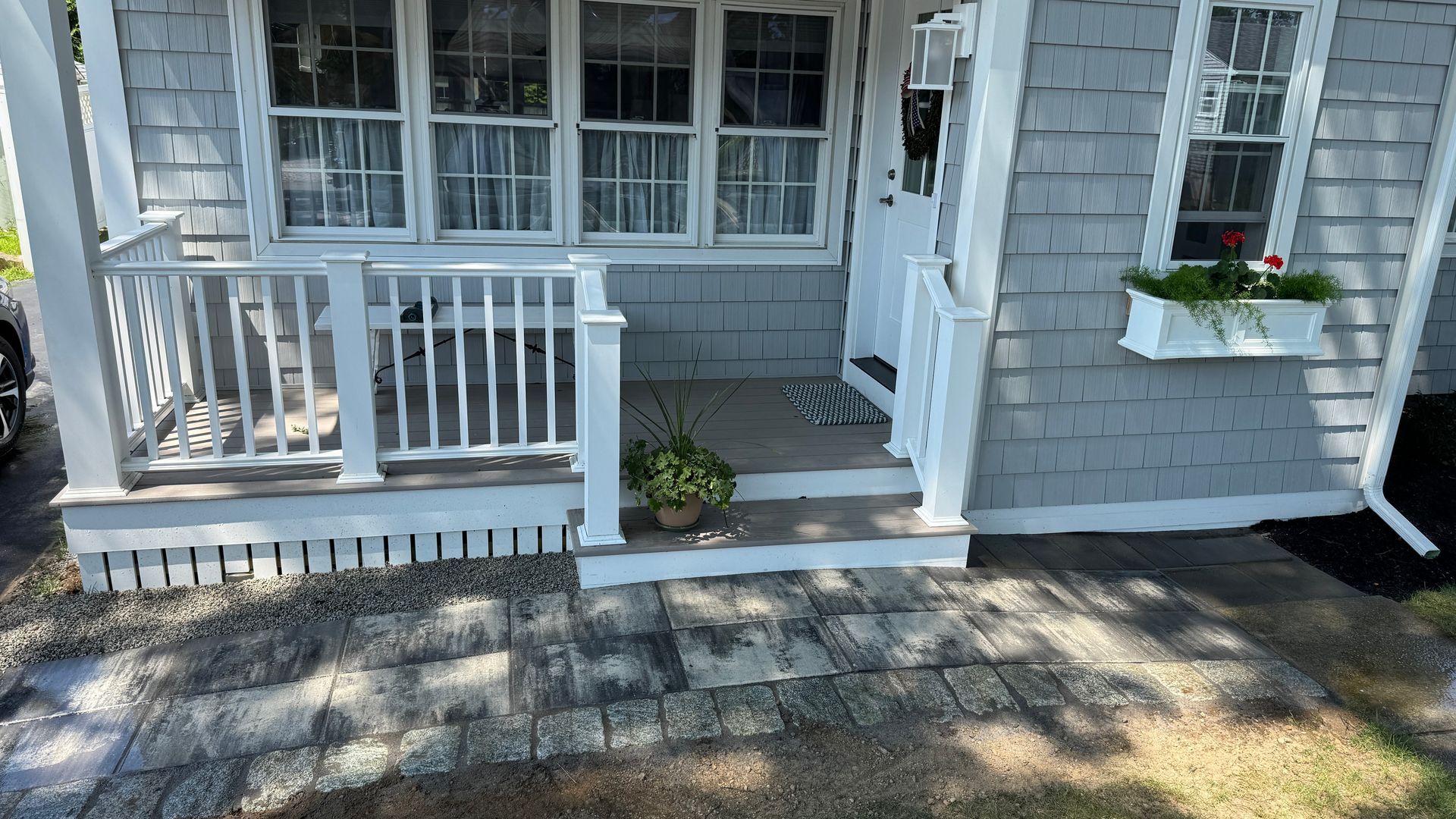 Gray shingled house with white porch and window flower box, brick walkway.