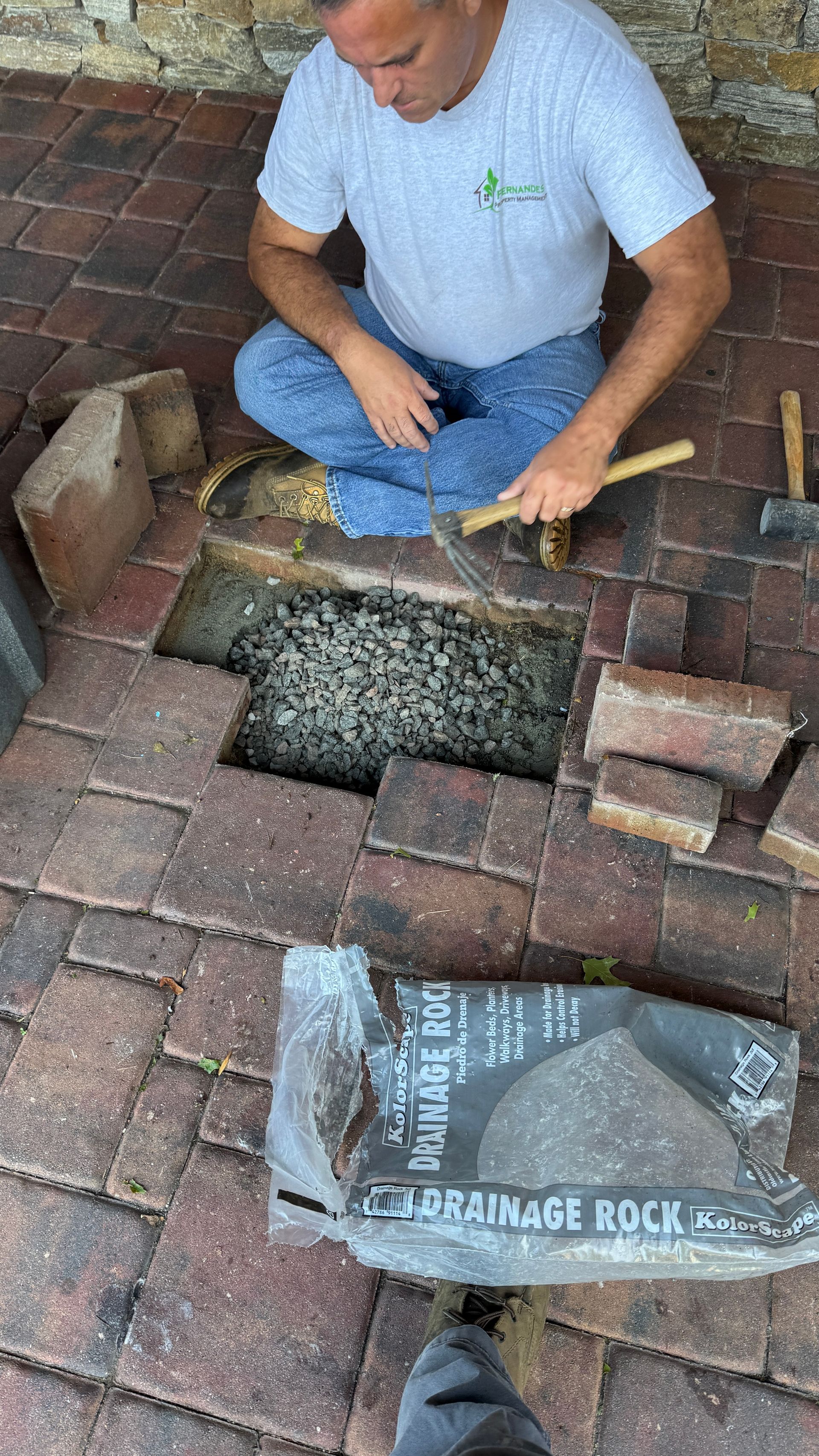 Man repairing brick patio, using hammer, surrounded by loose bricks and drainage rock.