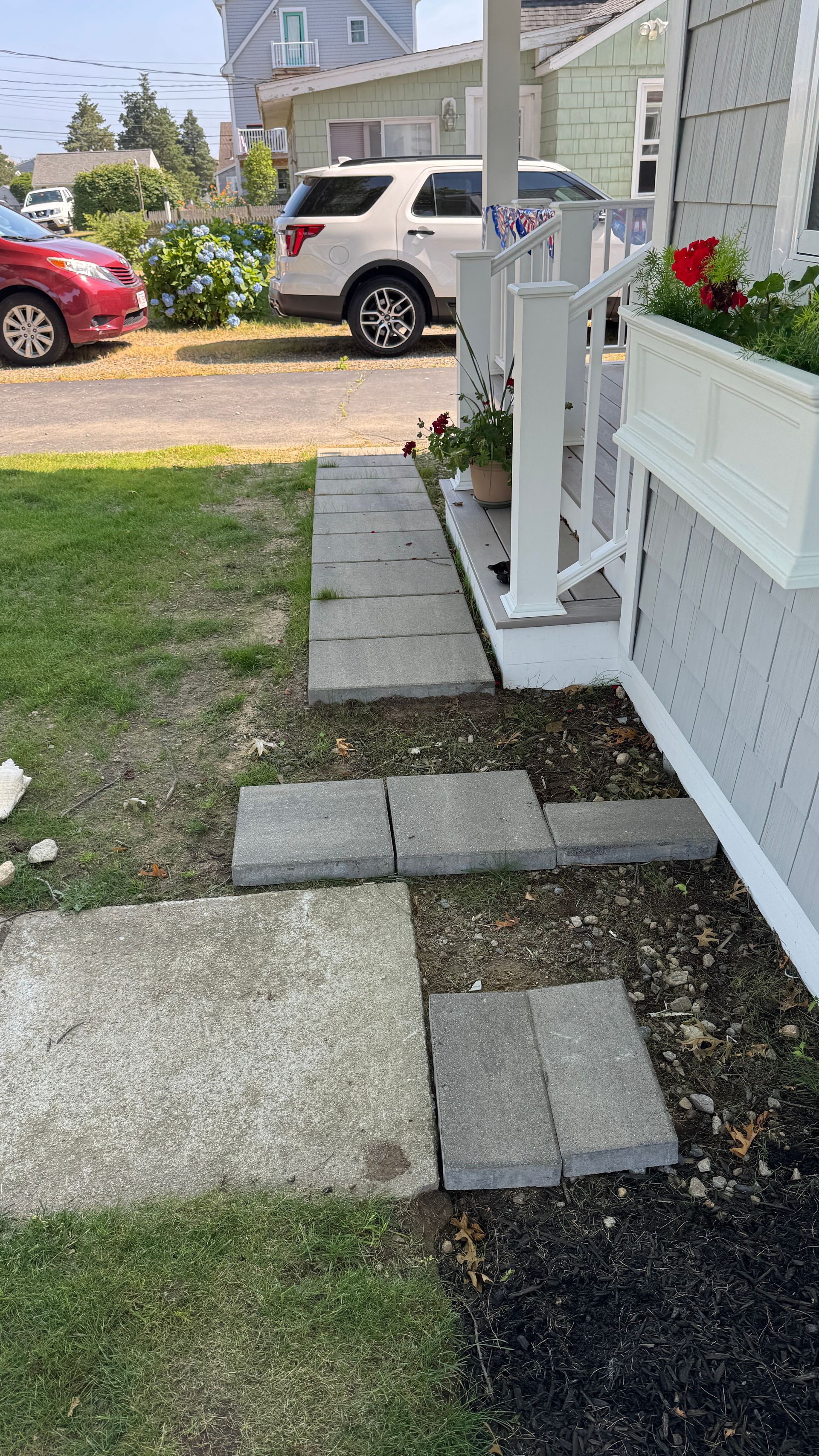 Stone walkway leads to a porch with white railing. Cars are parked in the background, near a house.