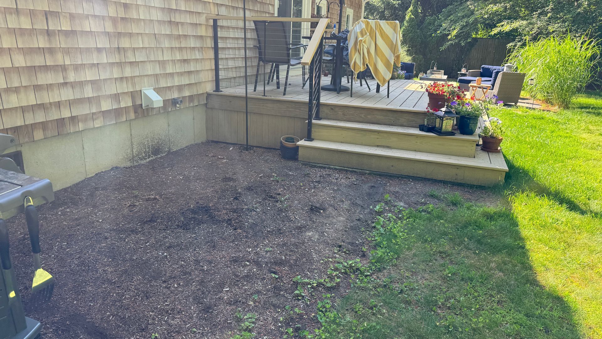 Gravel-covered area next to a wooden deck with steps and a grassy lawn, near a house with wood siding.