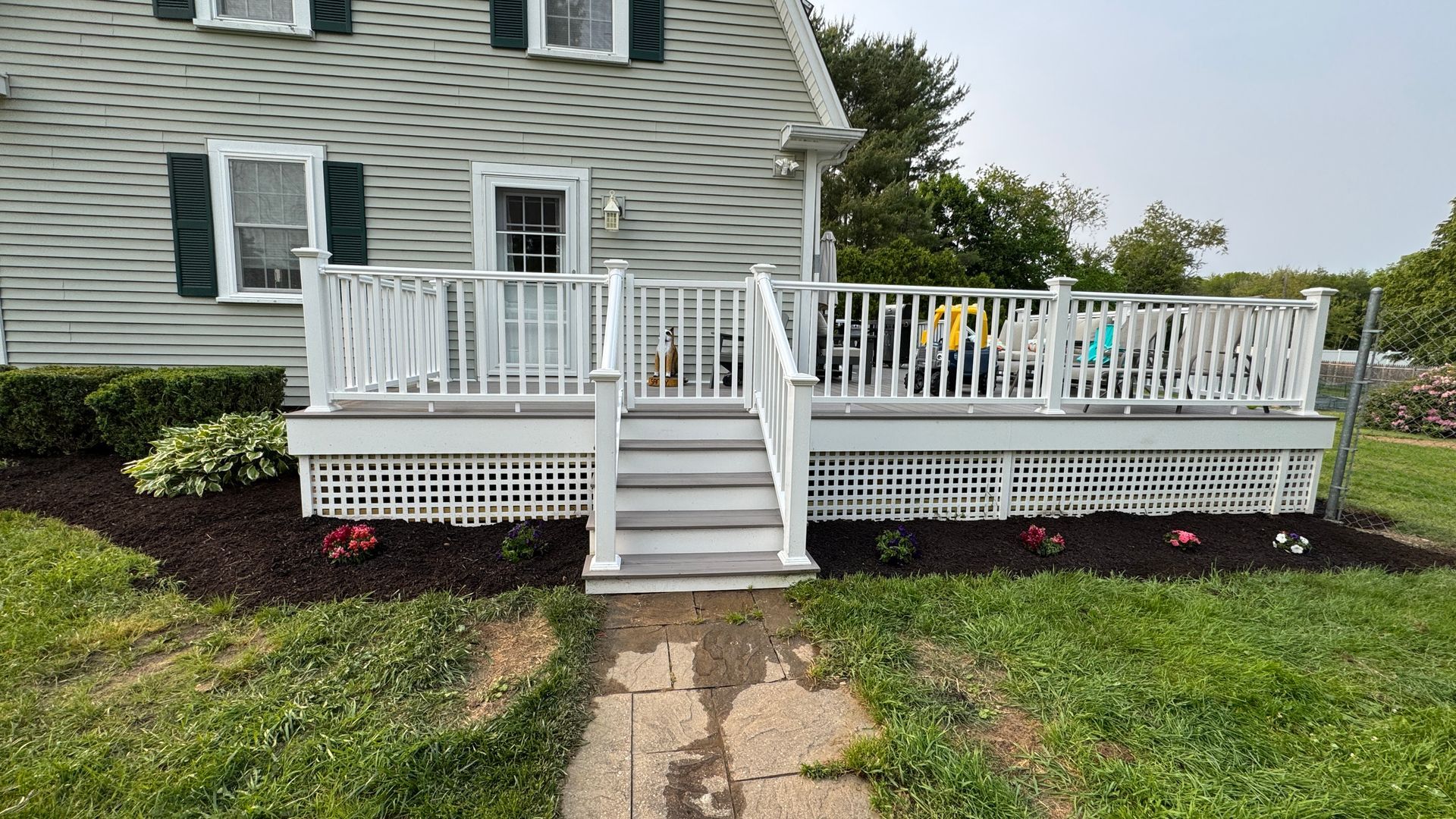 White deck with stairs in front of a light gray house. Landscaping with mulch and flowers.