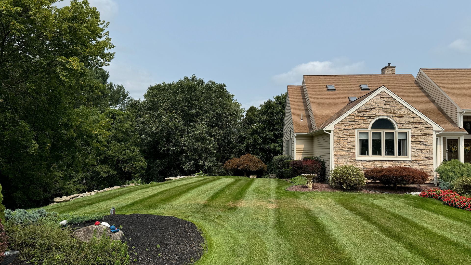 Lawn with striped grass in front of a stone house with a brown roof and a blue sky.