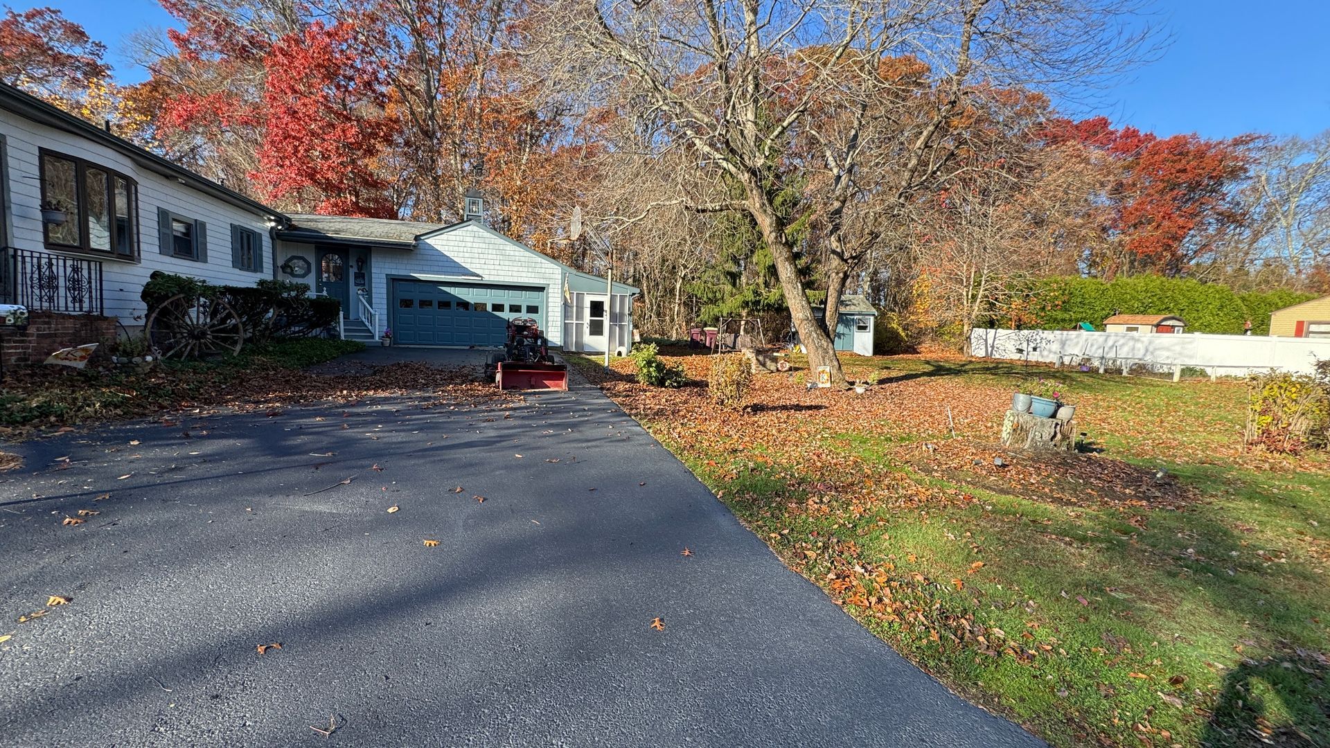 A house with a driveway lined with fallen leaves, trees with autumn colors, and a blue garage.