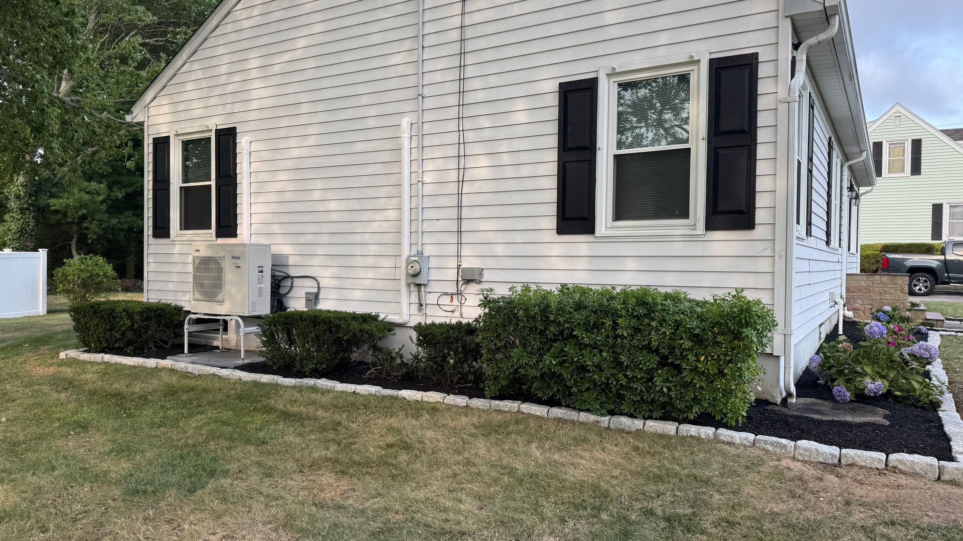 White house with black shutters, air conditioner, and bushes along a mulched bed.