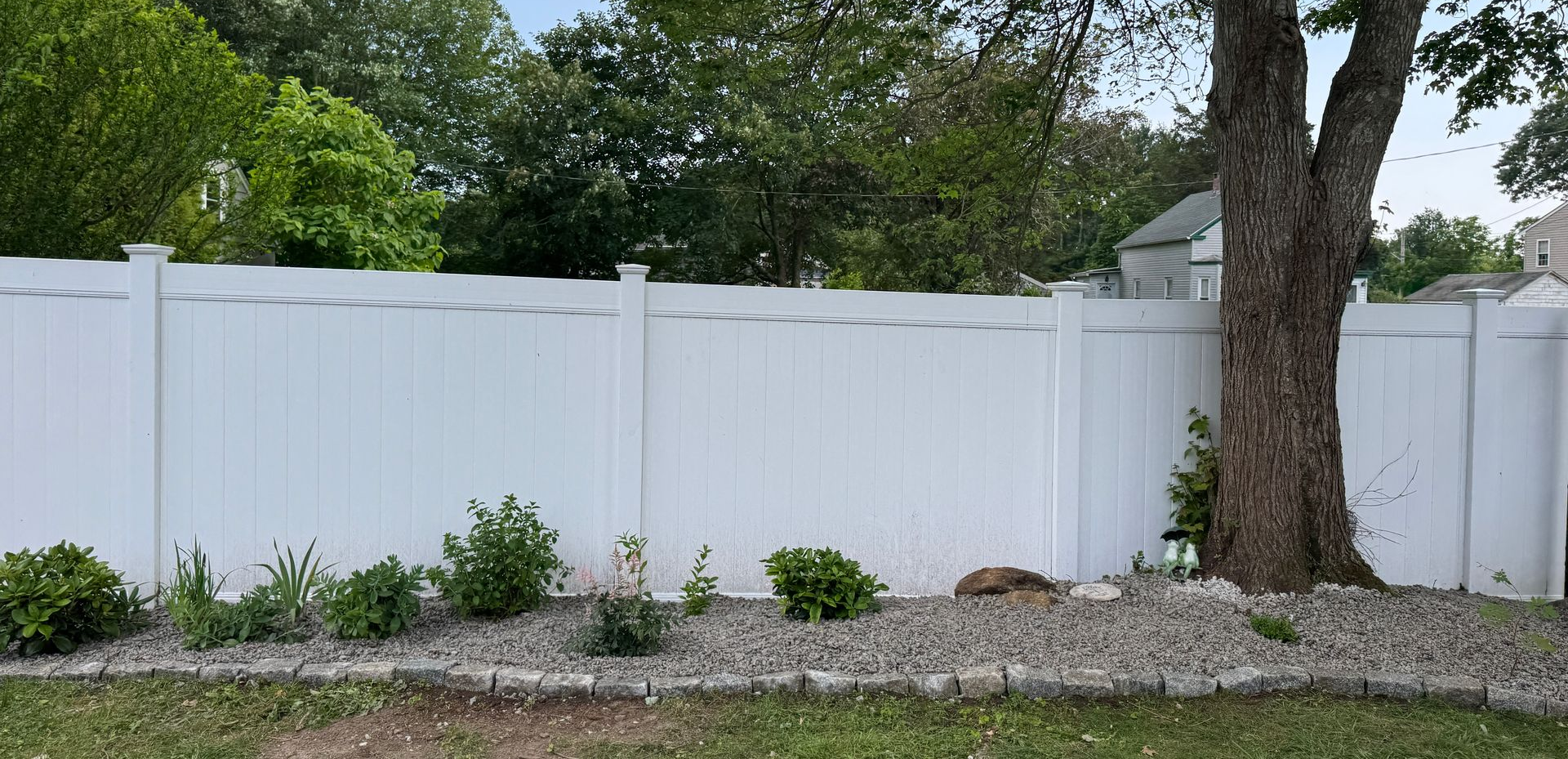 White fence with bushes in front, a tree to the right, and a grassy area in the foreground.