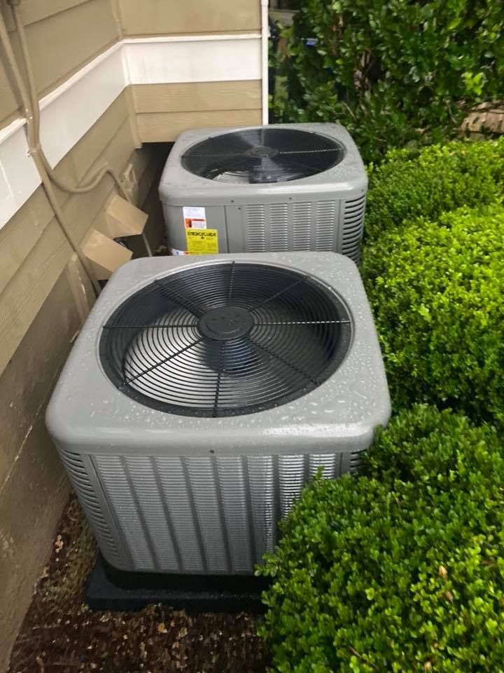 Two air conditioners are sitting next to each other on the side of a house.