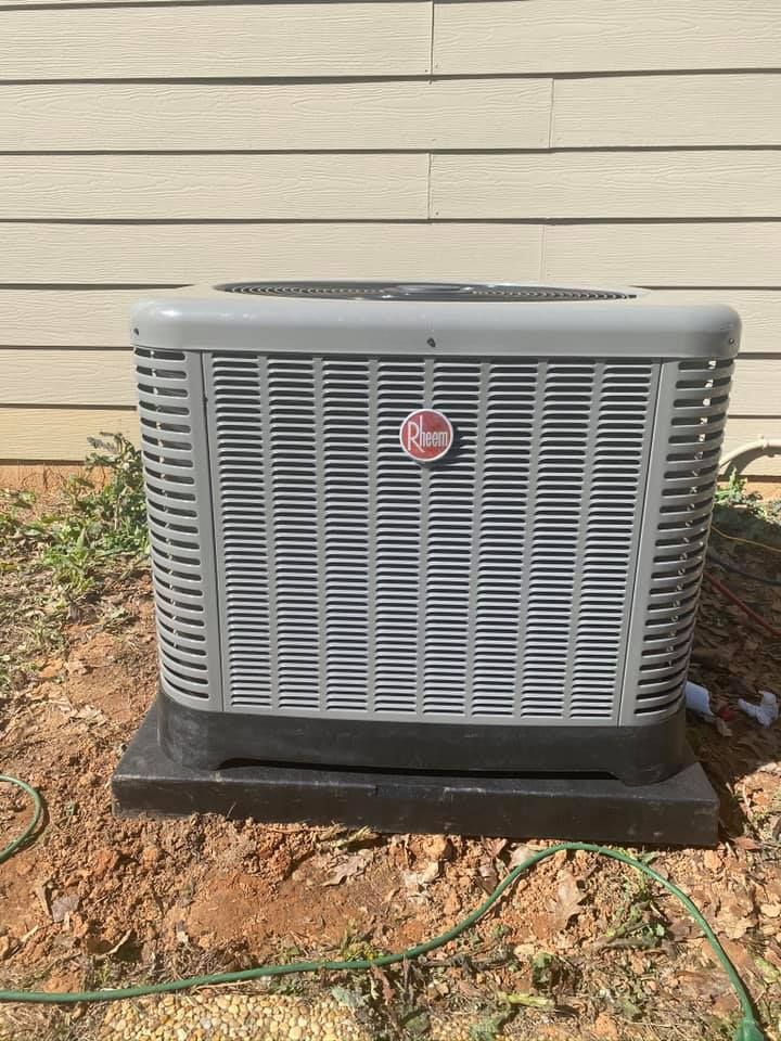 A gray air conditioner is sitting in the dirt in front of a house.