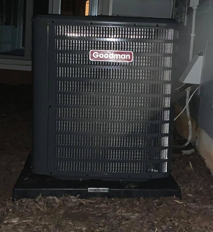 A goodman air conditioner is sitting on the ground outside of a house.