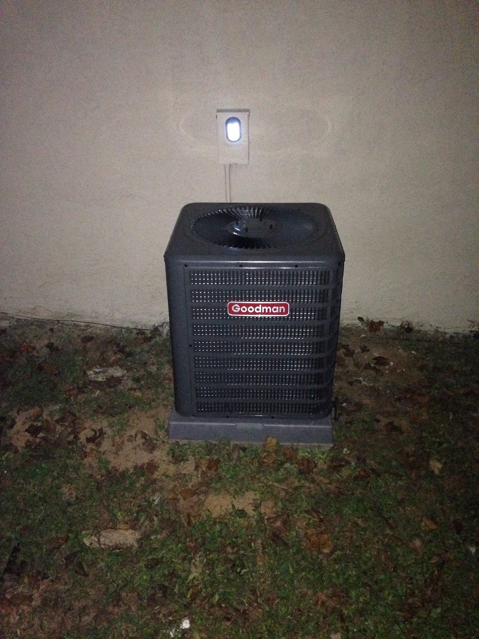 A black air conditioner is sitting in the grass next to a wall.