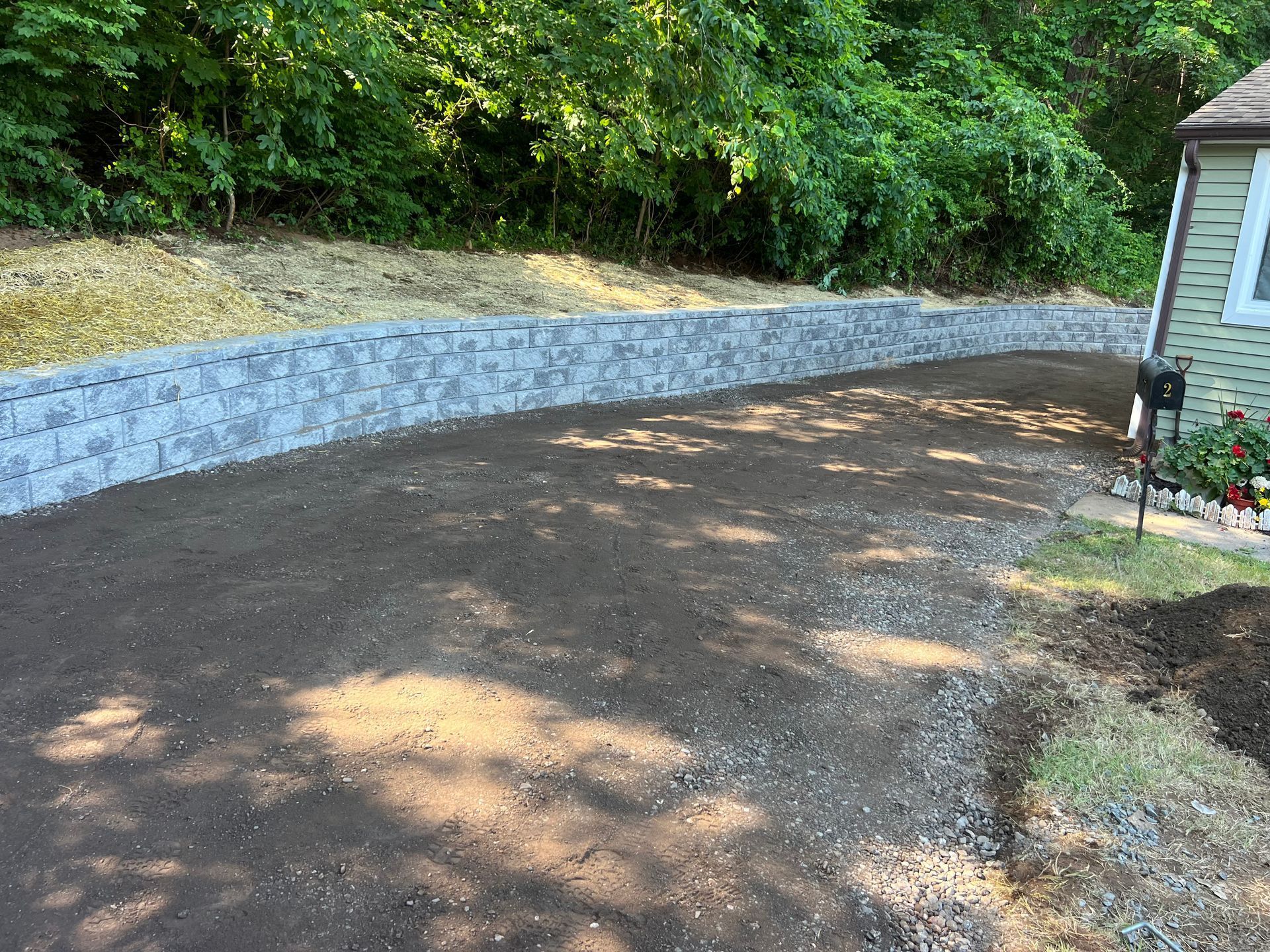 Gray retaining wall borders a gravel driveway, next to a house with trees in the background.