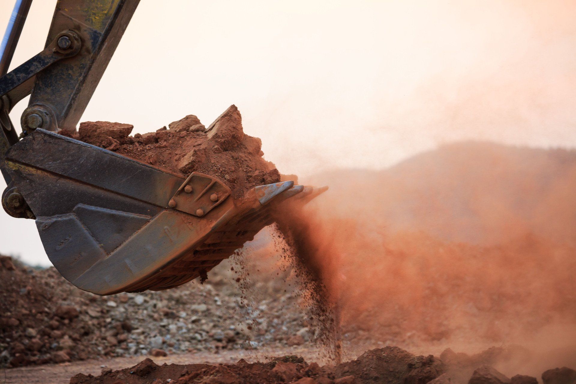Excavator bucket dumping reddish-brown soil, creating a cloud of dust.