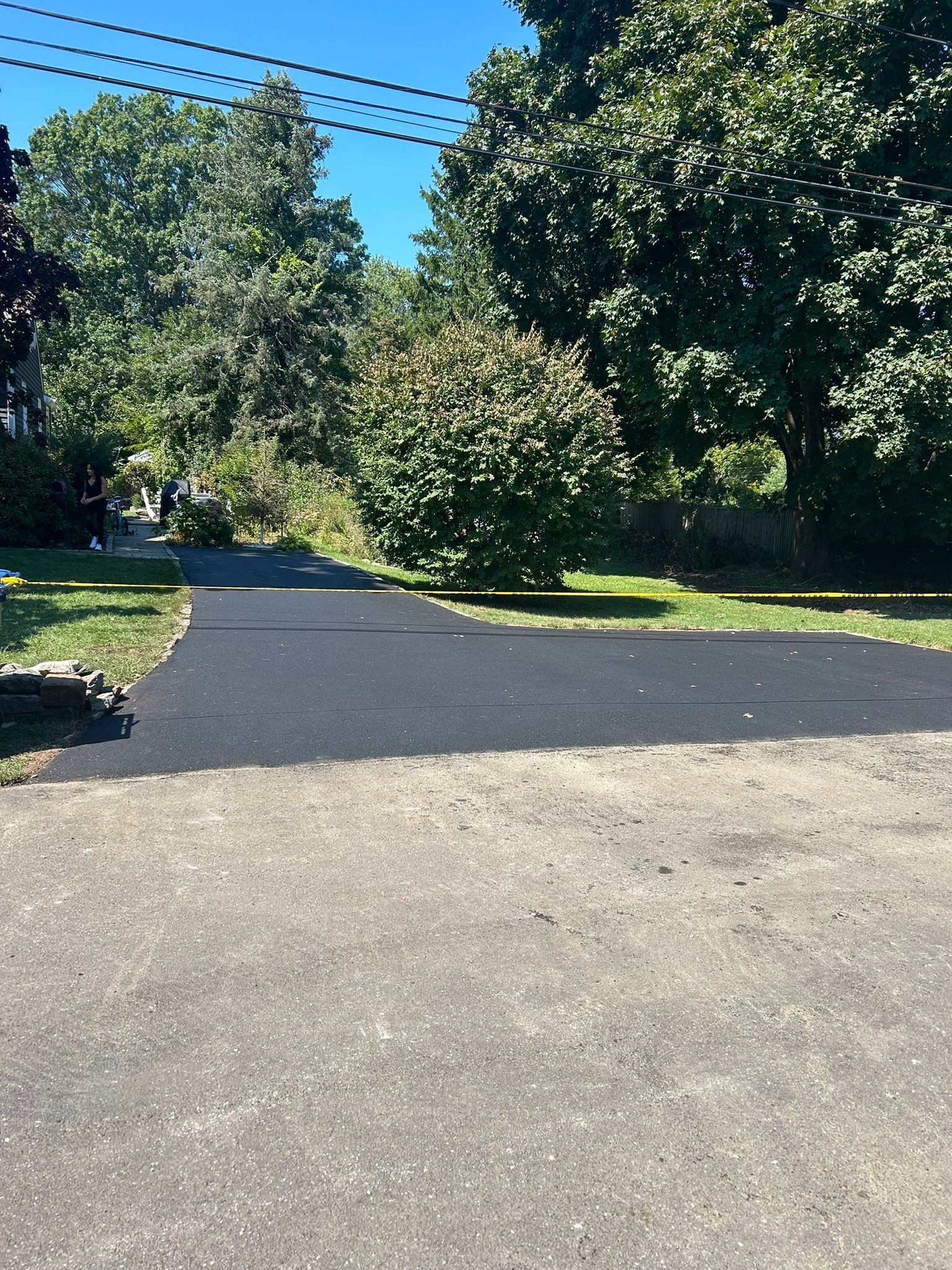 Asphalt driveway leading past a shrub and grassy area into a tree-lined setting on a sunny day.