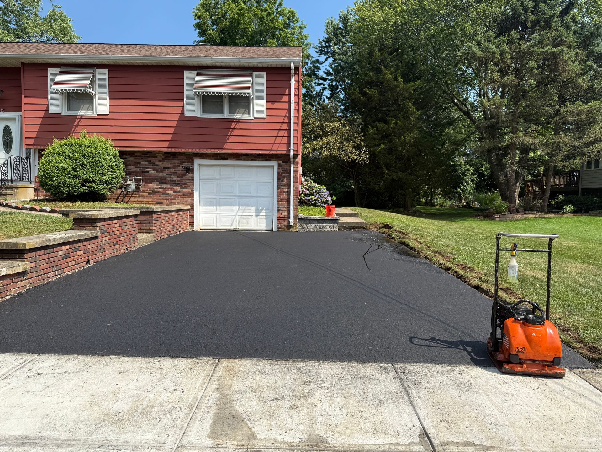 Newly paved black asphalt driveway in front of a red brick house with a white garage door. A vibratory plate is on the right.