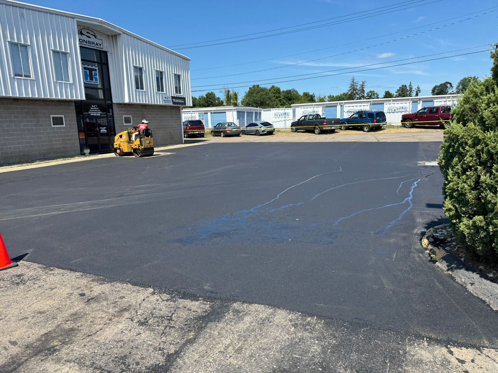 Newly paved parking lot with storage units and vehicles in the background. A worker operates a roller.