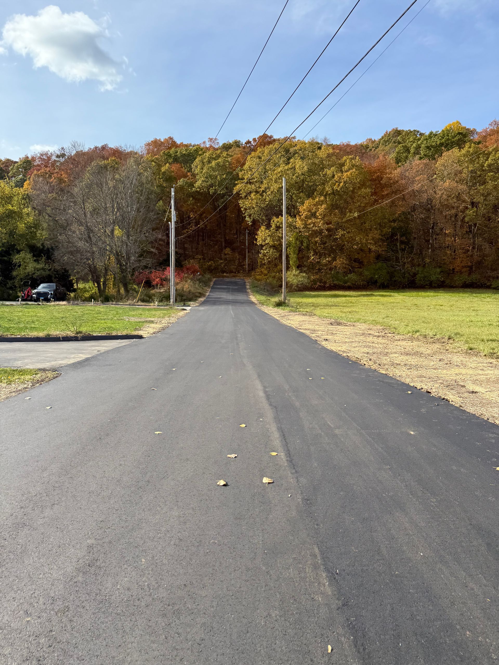 Paved road leading uphill towards a forest with vibrant fall foliage. Power lines overhead; a car parked to the side.