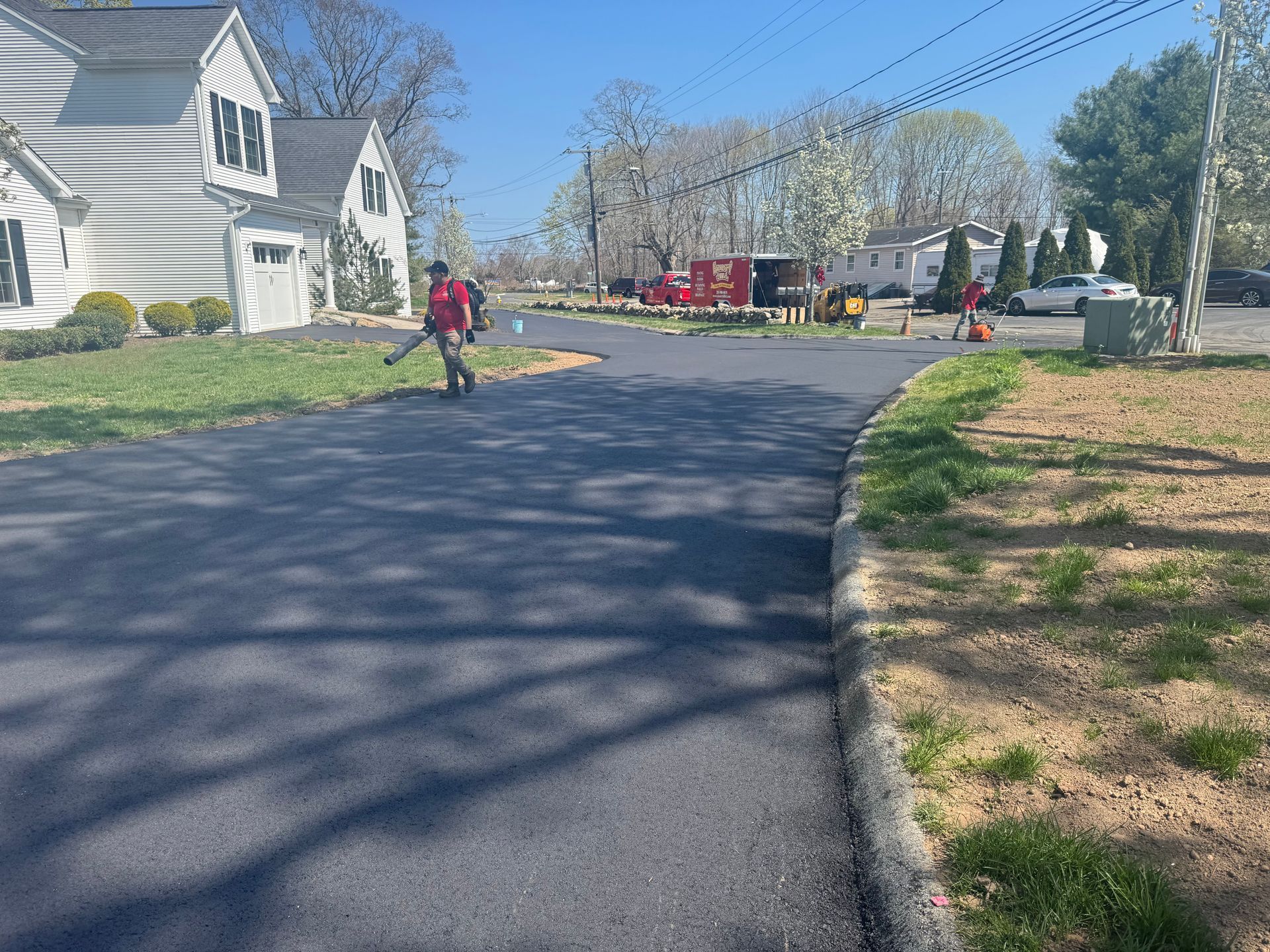 Man using leaf blower on a freshly paved road. Houses and equipment are in the background on a sunny day.