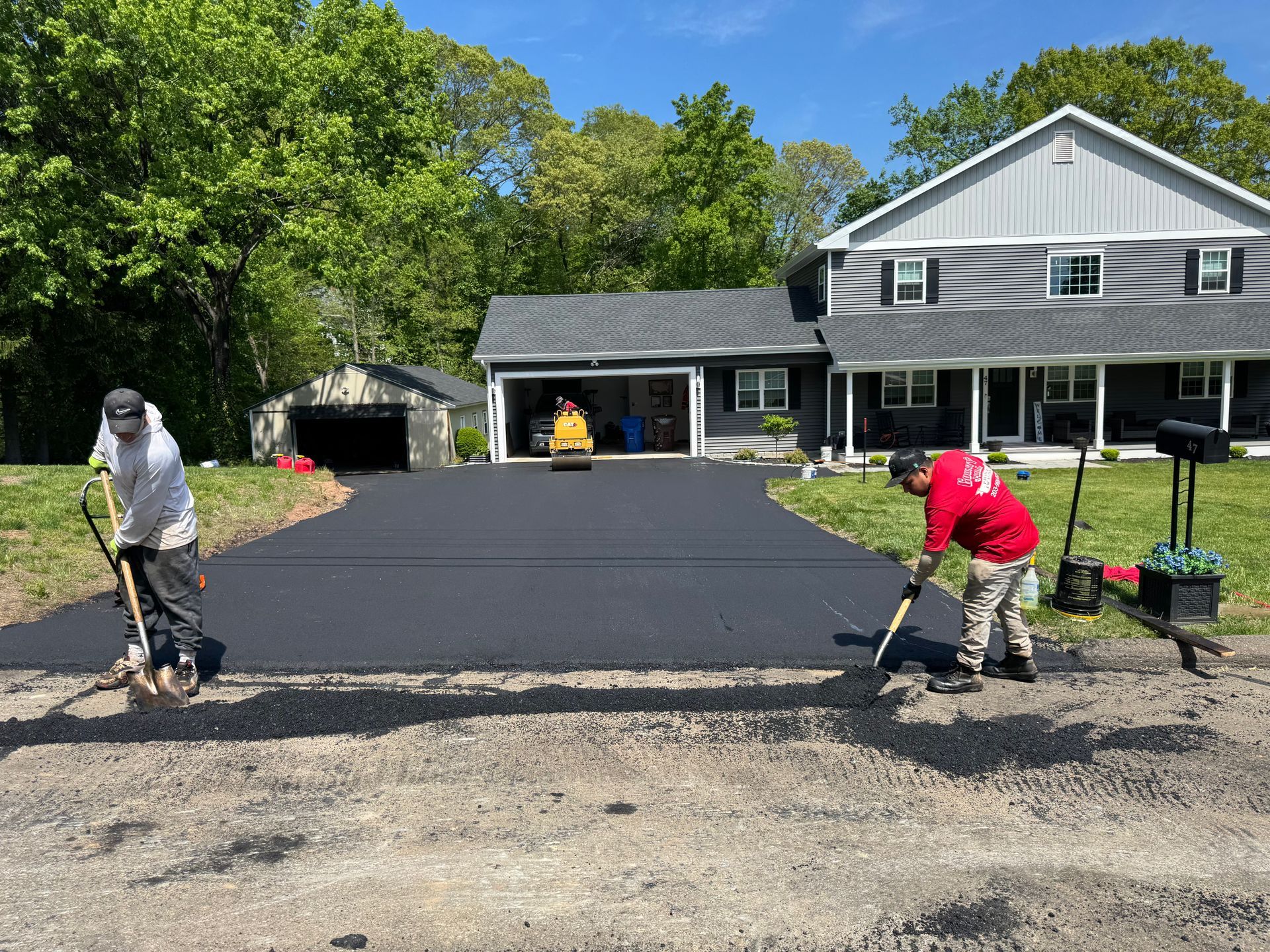 Workers paving a driveway in front of a gray house and garage on a sunny day.