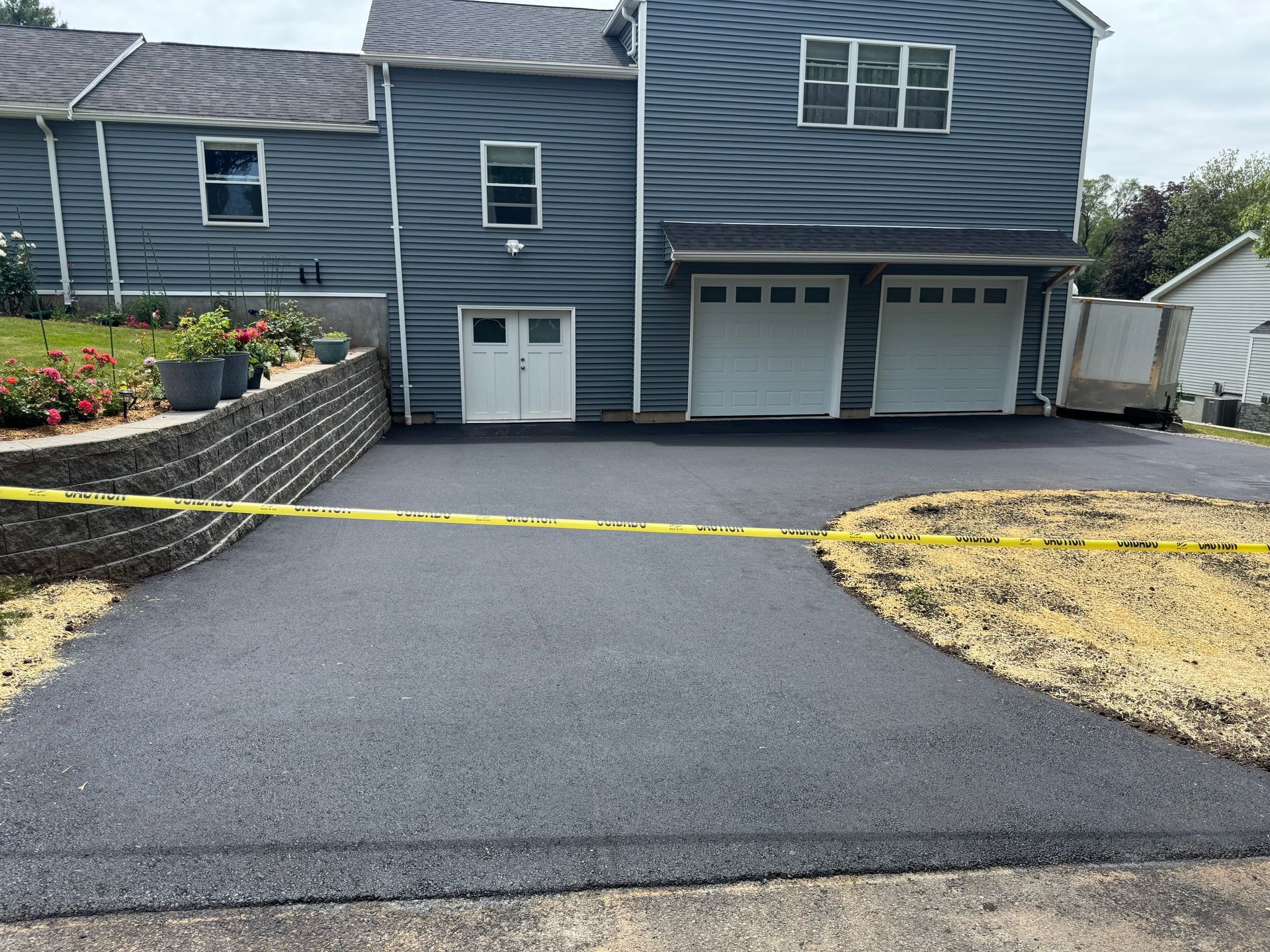 Newly paved asphalt driveway in front of a gray house with two garage doors. Yellow caution tape is present.
