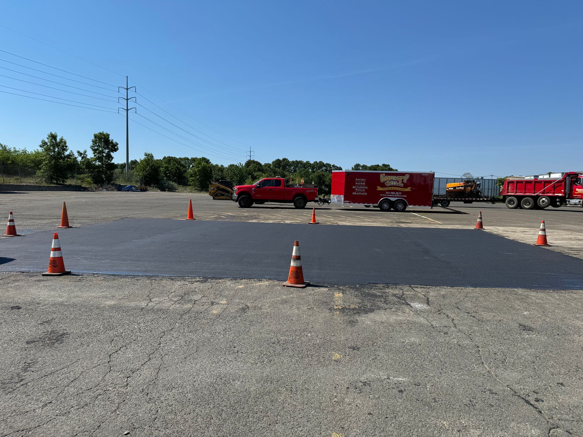Freshly paved asphalt patch, surrounded by orange traffic cones, construction vehicles in the background.