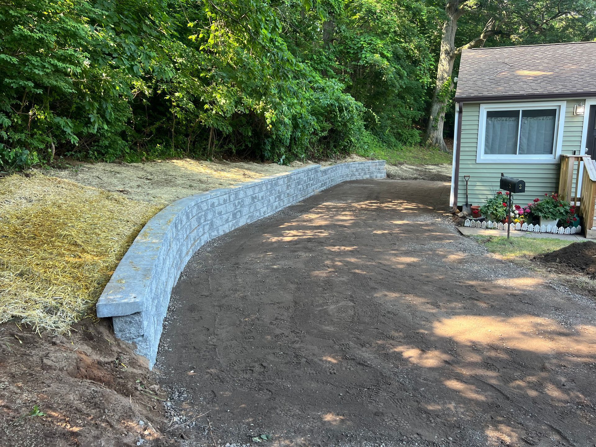 Curved stone retaining wall along a gravel driveway; a small house is in the background.