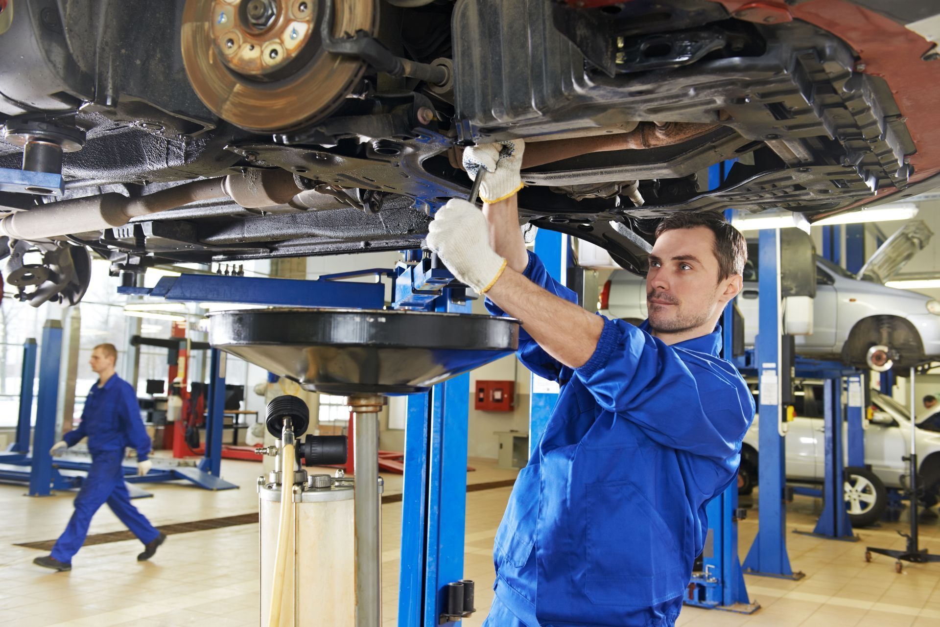 Mechanic working under a car on a lift in a garage, wearing blue coveralls and gloves.
