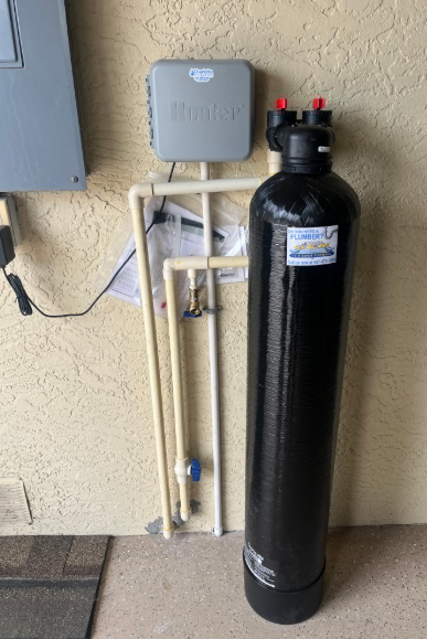 Black water softener tank with connected pipes and gray control box mounted on an exterior wall.