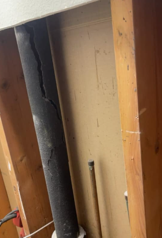 Damaged black insulation inside a wooden wall cavity with exposed wiring and beige backing panel