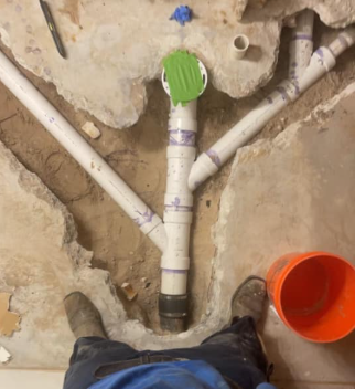 Plumbing pipes in a rough wall opening with a green cap and orange bucket on the floor.