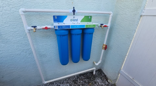 Blue water filtration system with three cartridges mounted on a white outdoor wall.
