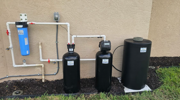 Water softener and filtration system with blue filter, two black tanks, and a black brine tank against an exterior wall.