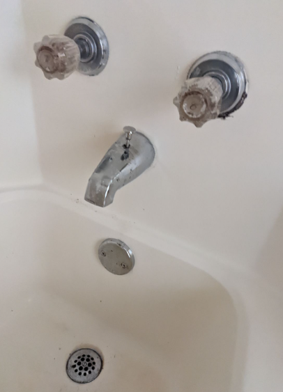 Bathtub faucet with two knobs and a silver spout above the drain, slightly stained white tub surround