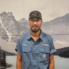 Man in blue work shirt and cap, standing in front of a mountain scene background.