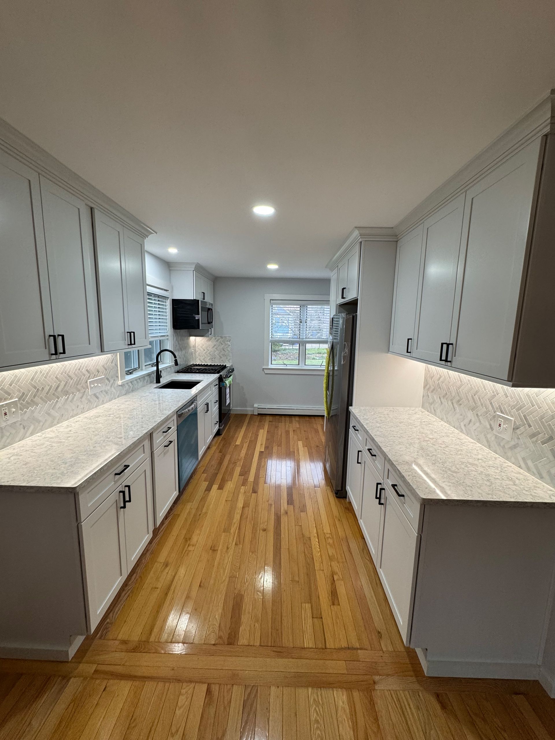 A long kitchen with white cabinets and hardwood floors.