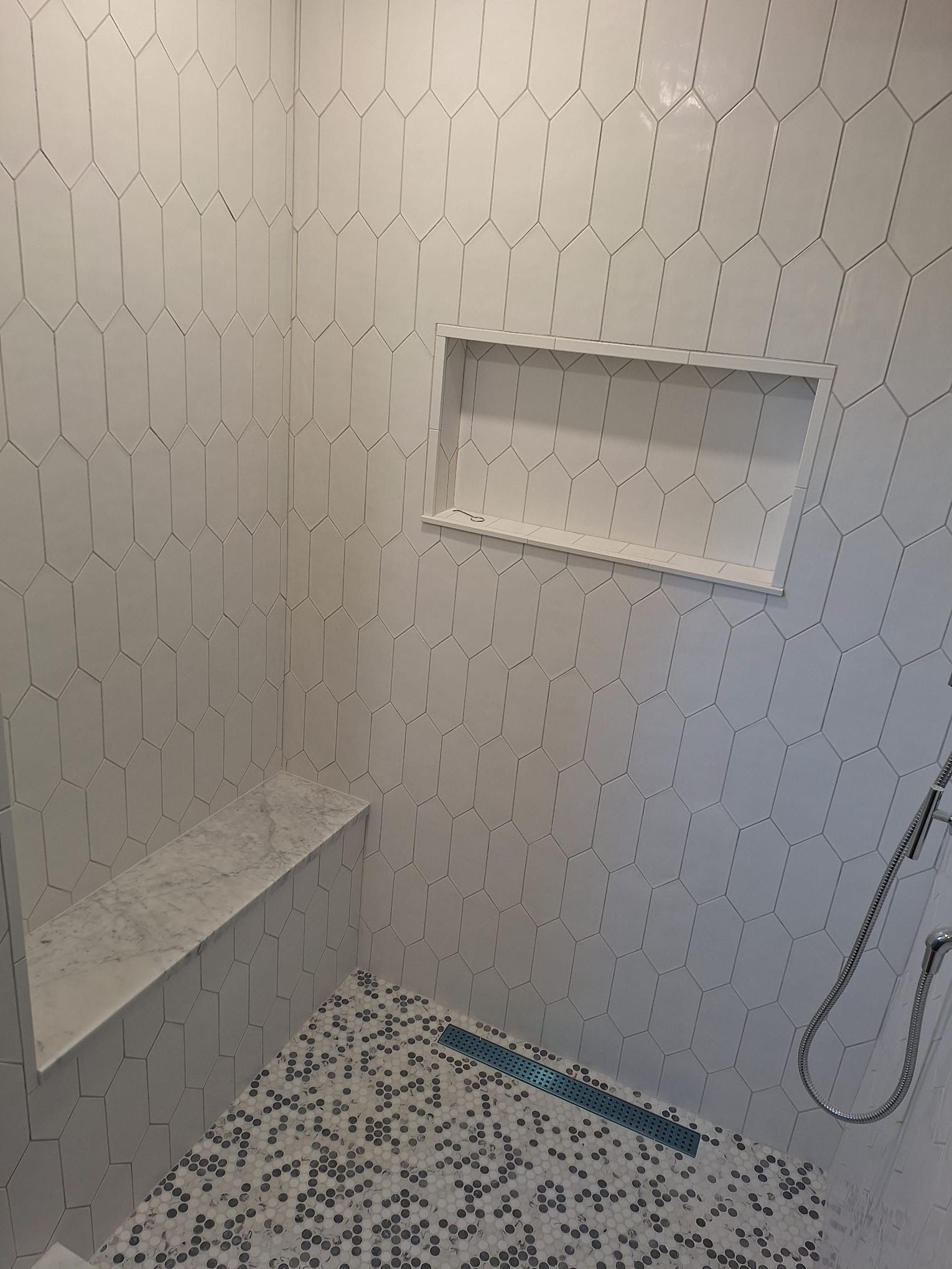 A shower with white tiles , a bench , and a shower head.