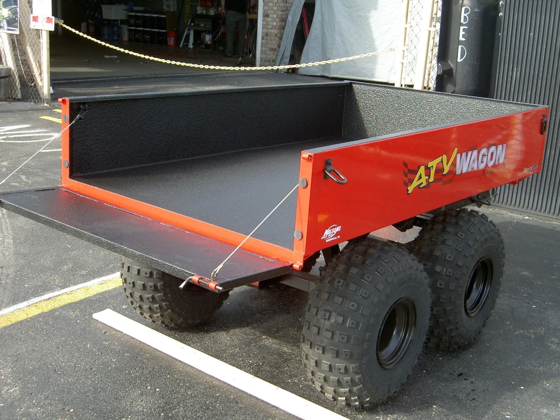 A red ATV Wagon trailer with thick off-road tires parked on pavement, featuring an open, drop-down tailgate.